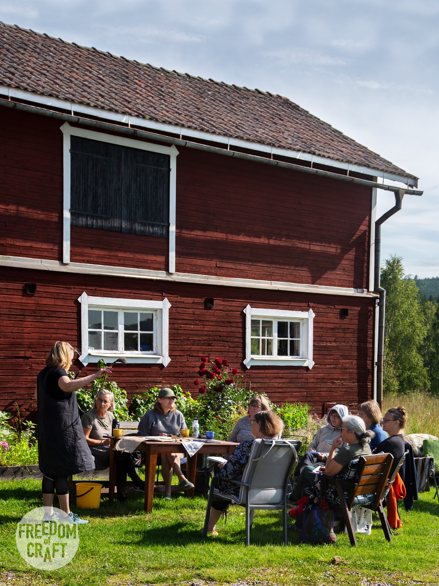 The studio at Böle textile farm. A traditional red barn, with white window linings. A group of students is sitting outside, listening to a teacher.